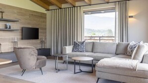Een moderne woonkamer met een bank, fauteuil en salontafel, een aan de muur bevestigde televisie en een groot raam met gordijnen.