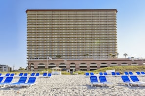 A high-rise hotel with multiple balconies, a beach area with sun loungers, and a clear sky.