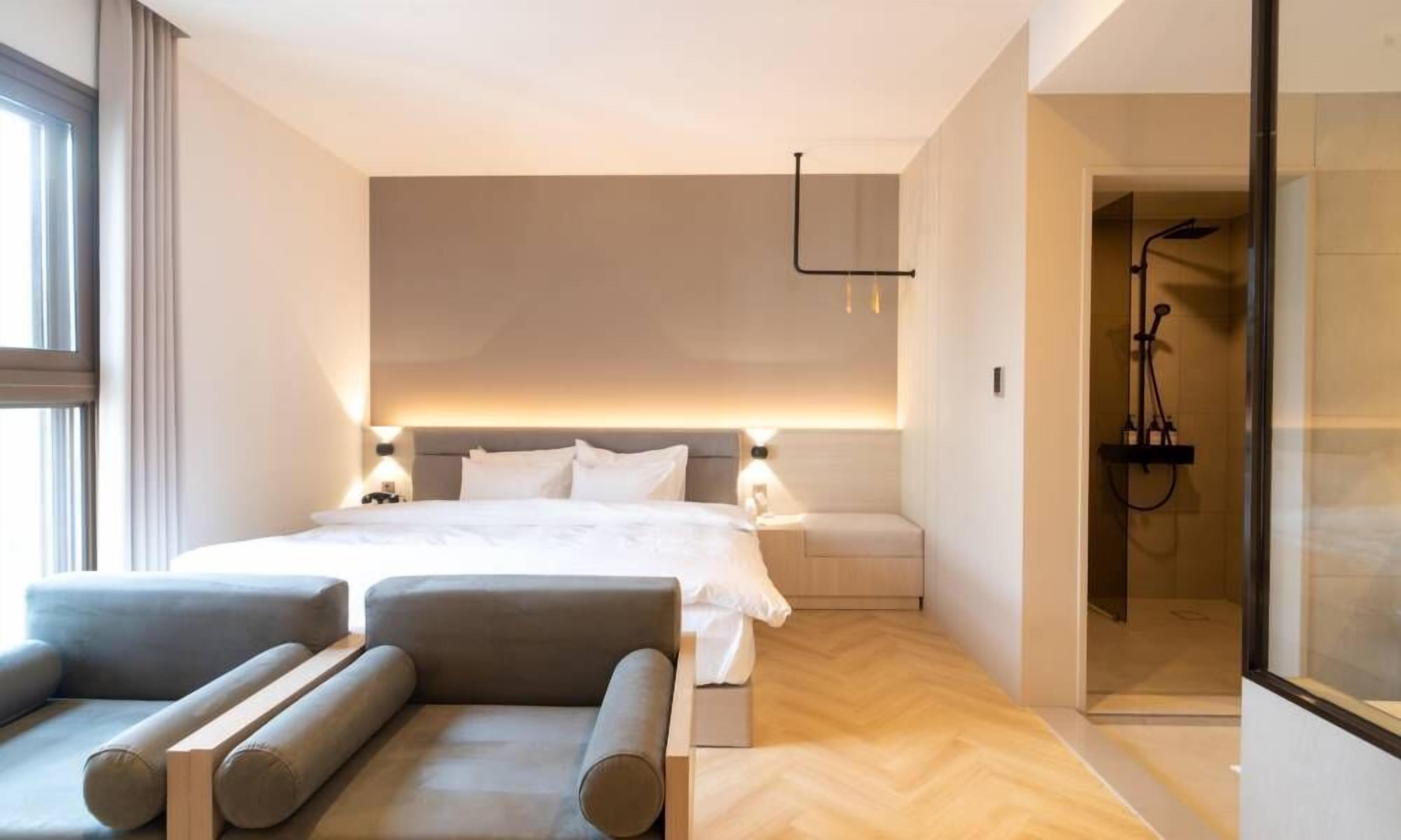 Una habitación de hotel moderna con una cama grande, zona de estar con sofás, cabina de ducha y puerta de vidrio que da a la ducha.