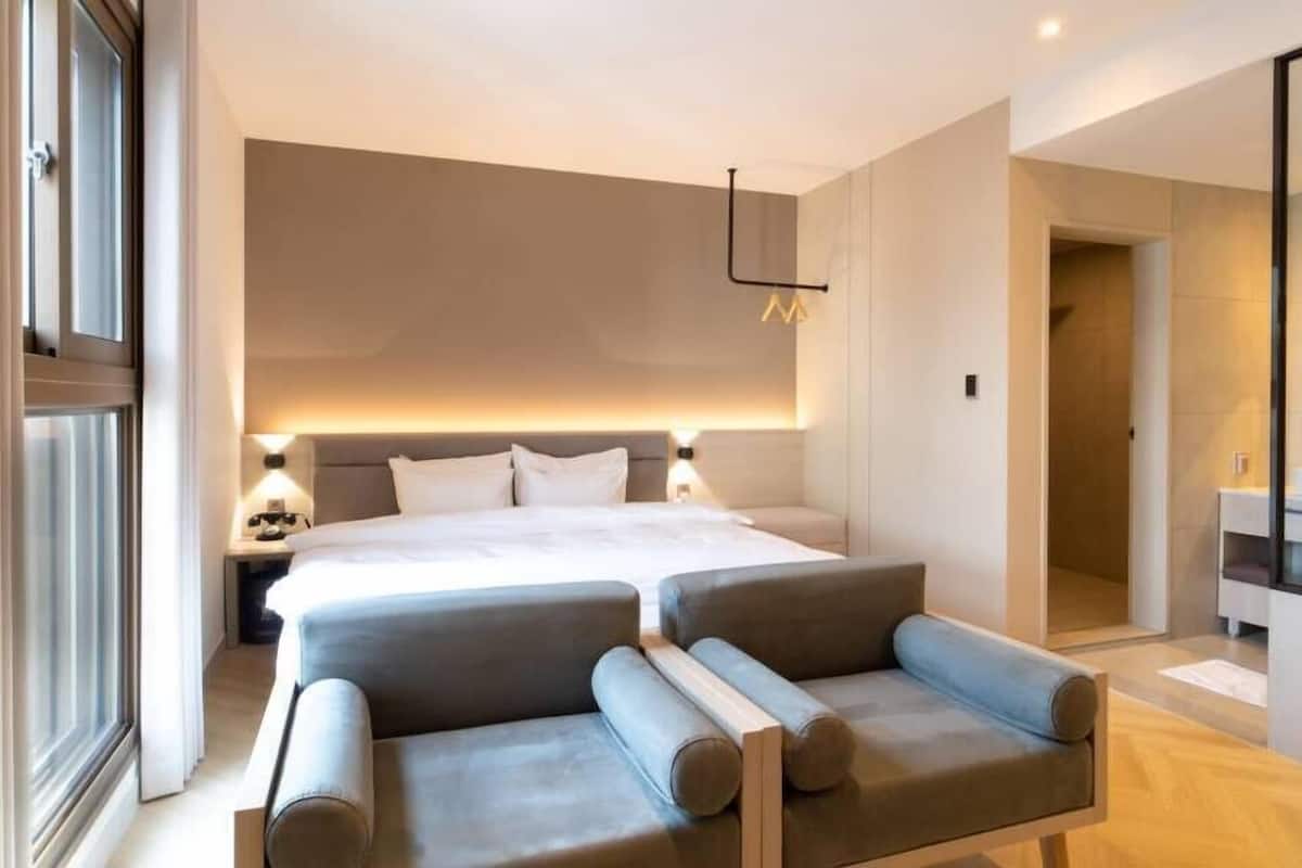 A modern hotel room with a large bed, two bedside tables, a sofa, and a bathroom visible through an open door.