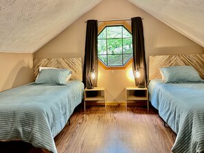 A cozy attic bedroom with two beds, a large window with curtains, and a wooden ceiling.
