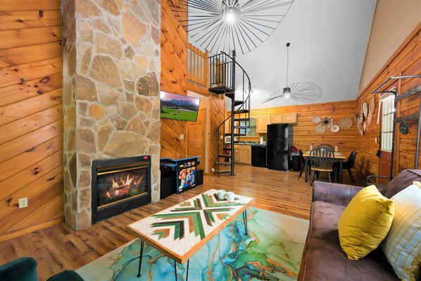 A cozy cabin interior with a stone fireplace, wooden walls, a spiral staircase, a dining area, and a TV mounted on the wall.