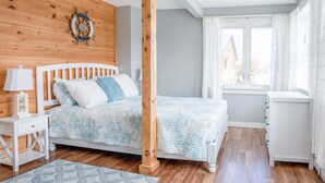 A bedroom with a wooden headboard, a bed with a blue quilt, a nightstand with a lamp, and a window with curtains. - Free Breakfast_In Downtown_Main Street Apt A (Bar Harbor)
