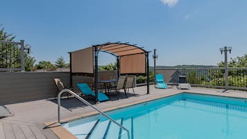 A swimming pool with a wooden deck, lounge chairs, a covered seating area, and a view of the landscape.