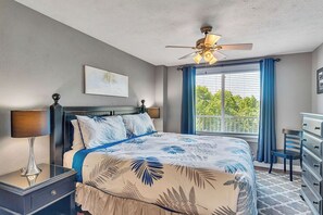 Une chambre à coucher comprenant un lit, une table de chevet, une commode, un ventilateur de plafond et une fenêtre avec des rideaux.