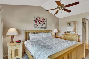 A bedroom with a bed, two bedside tables with lamps, a ceiling fan, and artwork on the walls.