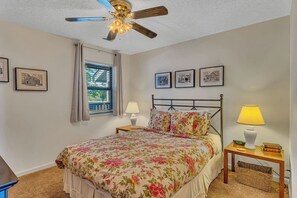 Une chambre à coucher avec un lit, un ventilateur de plafond, une fenêtre avec des rideaux, des lampes de chevet et des tableaux encadrés accrochés au mur.