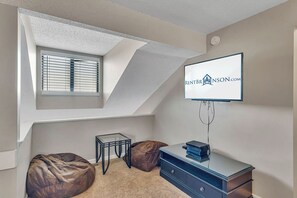 Une chambre avec un plafond en pente, un téléviseur fixé au mur, un bureau avec des tiroirs et une fenêtre équipée de stores.