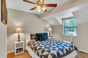 Une chambre avec un plafond en pente, un lit recouvert d’une couvre-lit à motifs géométriques, deux tables de chevet avec des lampes, une fenêtre avec des stores et un ventilateur de plafond.