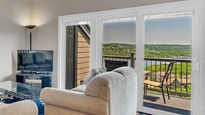 Een woonkamer met een televisie, bank en een balkon met uitzicht op een groen landschap.