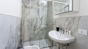 Une salle de bain moderne avec une douche à l’italienne en verre, un lavabo blanc et des murs en marbre.