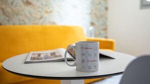 Une tasse blanche ornée d’icônes liées au voyage, posée sur une petite table ronde, avec une chaise jaune en arrière-plan.