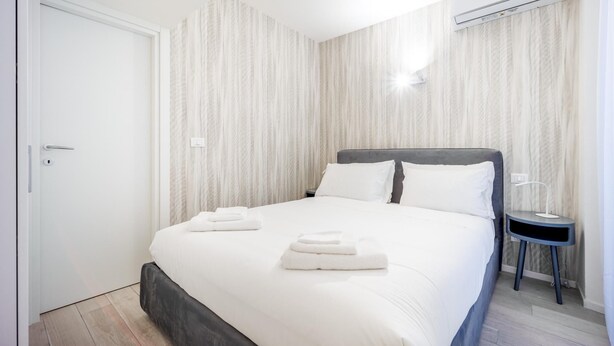 A neatly made bed with two folded towels, a bedside table with a lamp, and a wardrobe with a door. - Italianway - The B Village Standard - 03 (Milan)