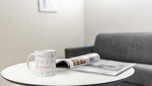 A white mug with Italianway branding on a small round table, a magazine, and a grey sofa in the background. - Italianway - The B Village Standard - 03 (Milan)