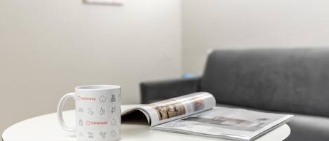 Una taza blanca con la marca Italianway sobre una mesita redonda, una revista y un sofá gris al fondo.
