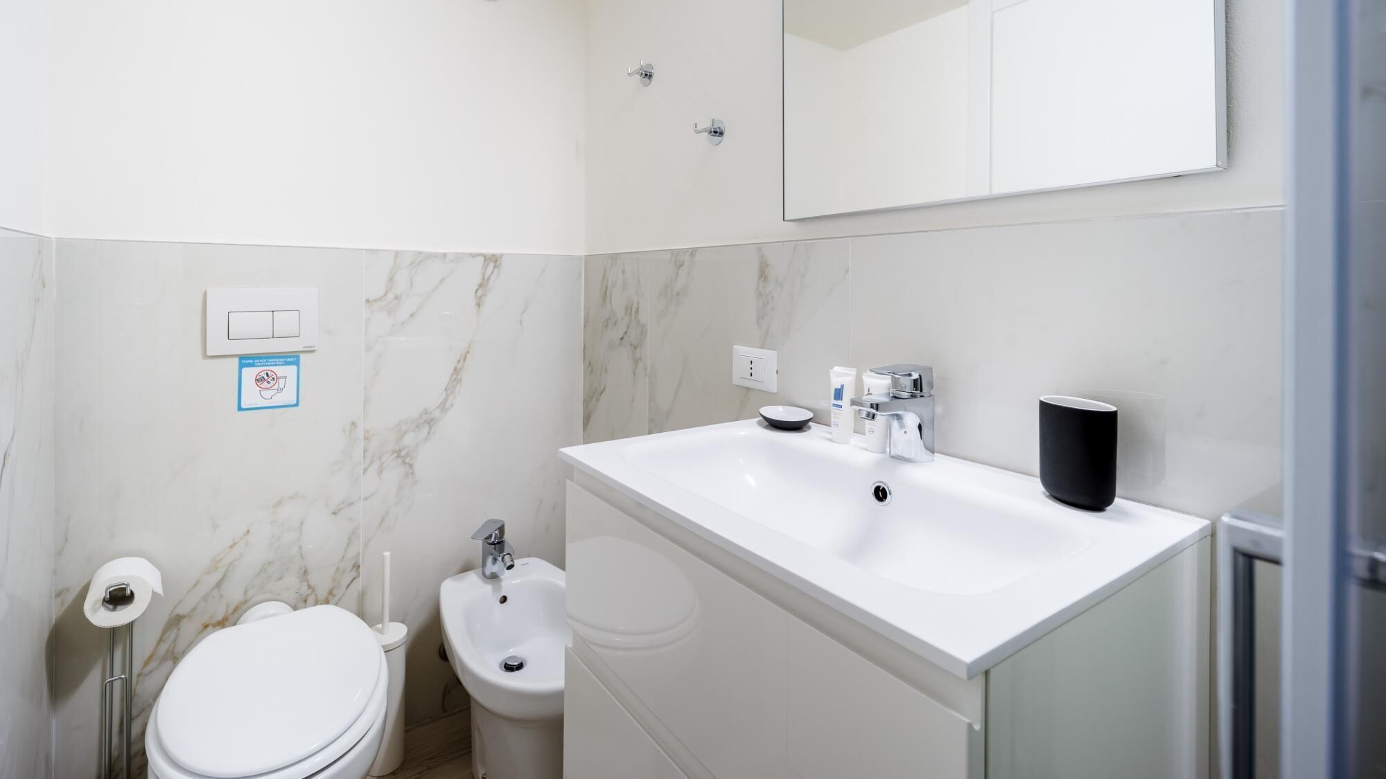 Een moderne badkamer met een wit wastafel, bidet, toilet en een spiegel boven het wastafel.