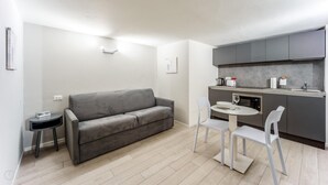 A compact living space with a grey sofa, a small round table, and two white chairs. A kitchenette with grey cabinets and a countertop is visible. - Italianway - The B Village Standard - 03 (Milan)