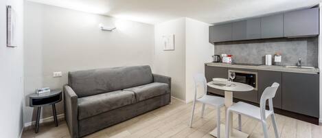 Un espacio habitable compacto con un sofá gris, una mesita redonda pequeña y dos sillas blancas. Se ve una kitchenette con armarios grises y una encimera.