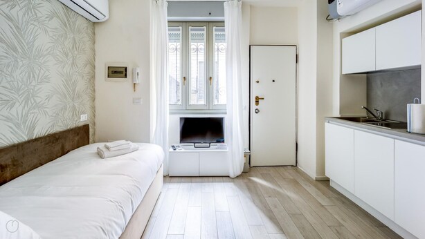 A compact hotel room with a bed, a small TV, a kitchenette, and a window with a view. - Italianway - The B Village Standard - 02 (Milan)
