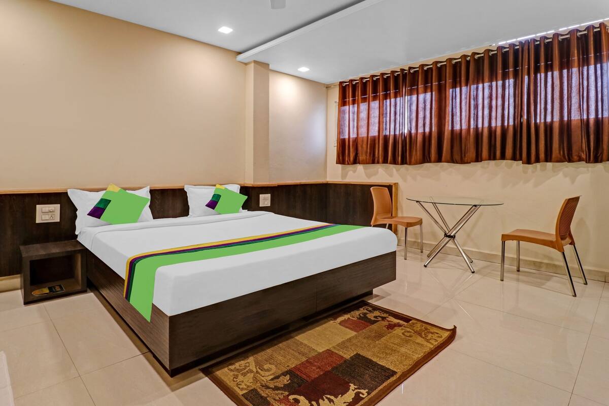 Een hotelkamer met een groot bed, een kleintje tafeltje, twee stoelen, een nachtkastje en een raam met gordijnen.