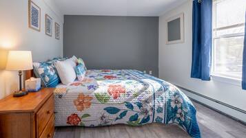Une chambre à coucher avec une couvre-lit à motifs floraux, une table de chevet en bois avec une lampe et une fenêtre ornée de rideaux bleus.
