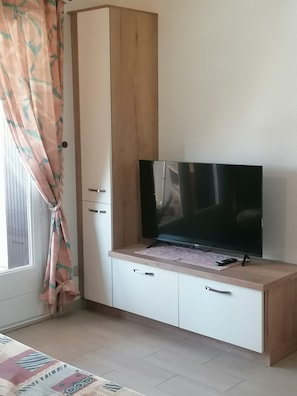 A room with a TV on a wooden cabinet, a window with curtains, and a patterned rug. - Apartment in quiet residence near the sea (Leucate)