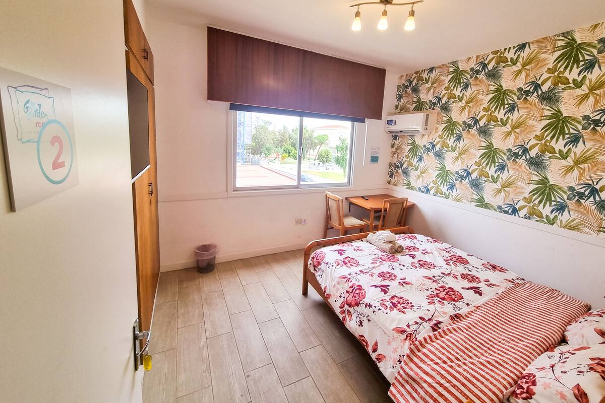 A bedroom with a bed, a small table, and a chair. There is a window with a view of the outside, a wall-mounted air conditioner, and a door with a lock.