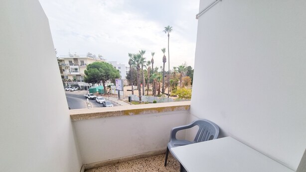 Un balcón con una mesa y dos sillas, piso de baldosas y vista a la calle con coches y palmeras.