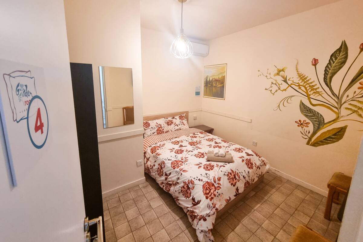 A bedroom with a bed, a mirror, a ceiling light, a wall-mounted air conditioner, and a floral mural.