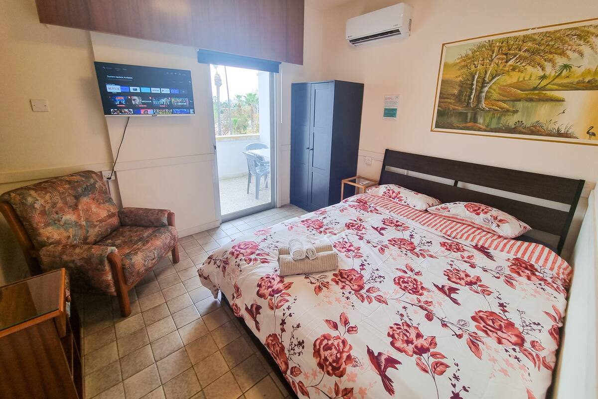 A hotel room with a bed, a chair, a TV mounted on the wall, an air conditioner, and a painting on the wall.