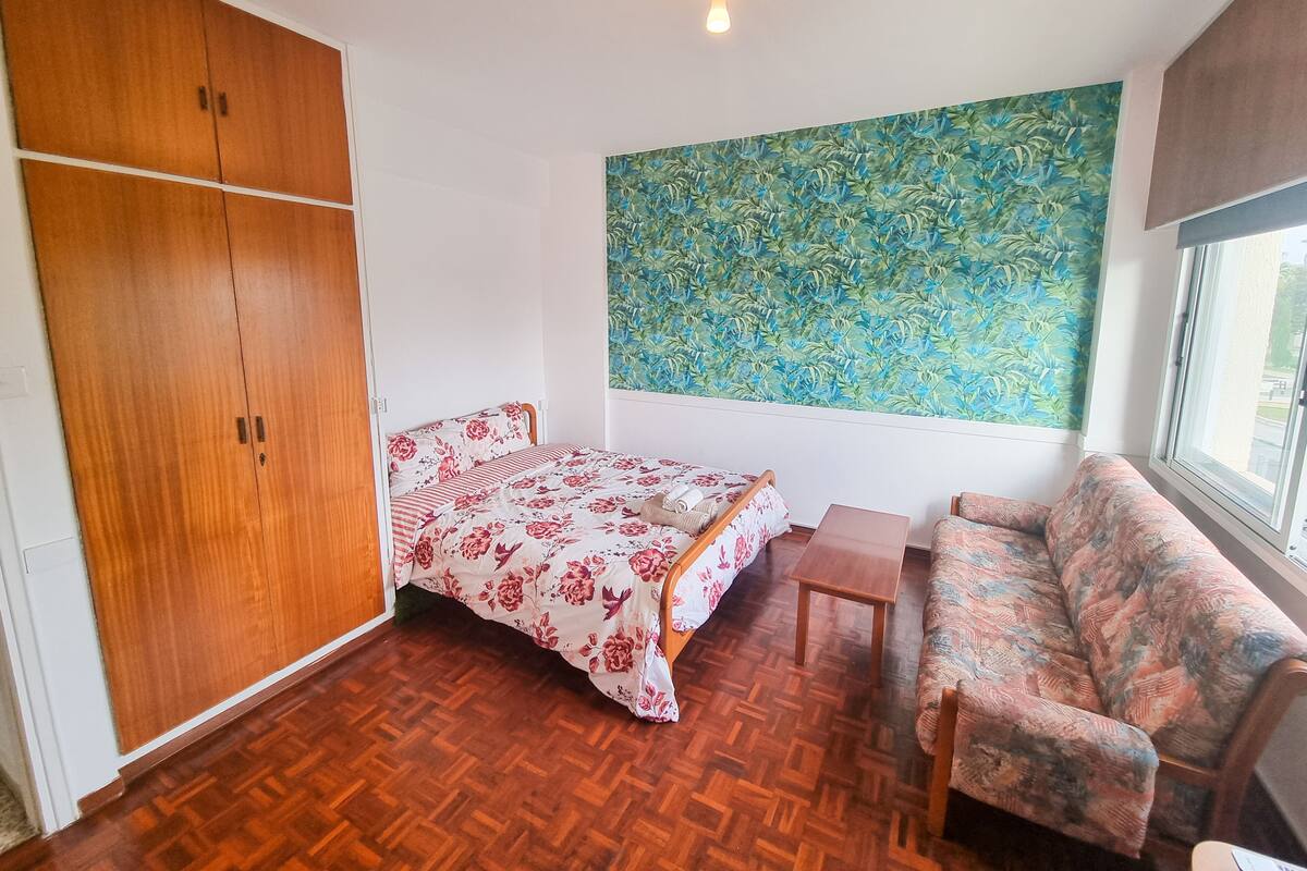 A room with a bed, a sofa, a wooden wardrobe, and a large floral-patterned wall hanging.