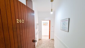 Un pasillo con una puerta de madera identificada con el número “202”, suelo de baldosas, una luz en el techo y un cartel de bienvenida en la pared.