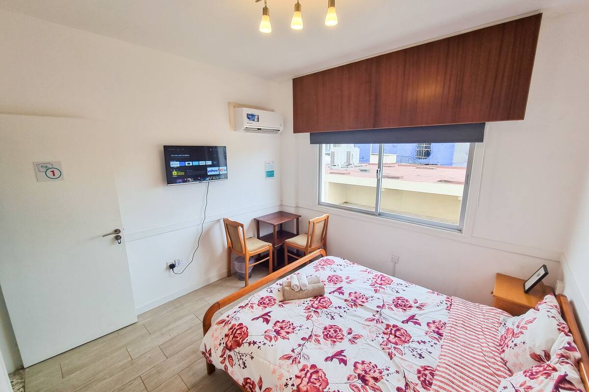 A bedroom with a bed, a desk, a chair, a TV mounted on the wall, and a window with a view of buildings outside.