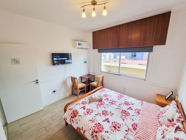 A bedroom with a bed, a desk, a chair, a TV mounted on the wall, and a window with a view of buildings outside. - Gardenrooms Larnaca (Larnaca)