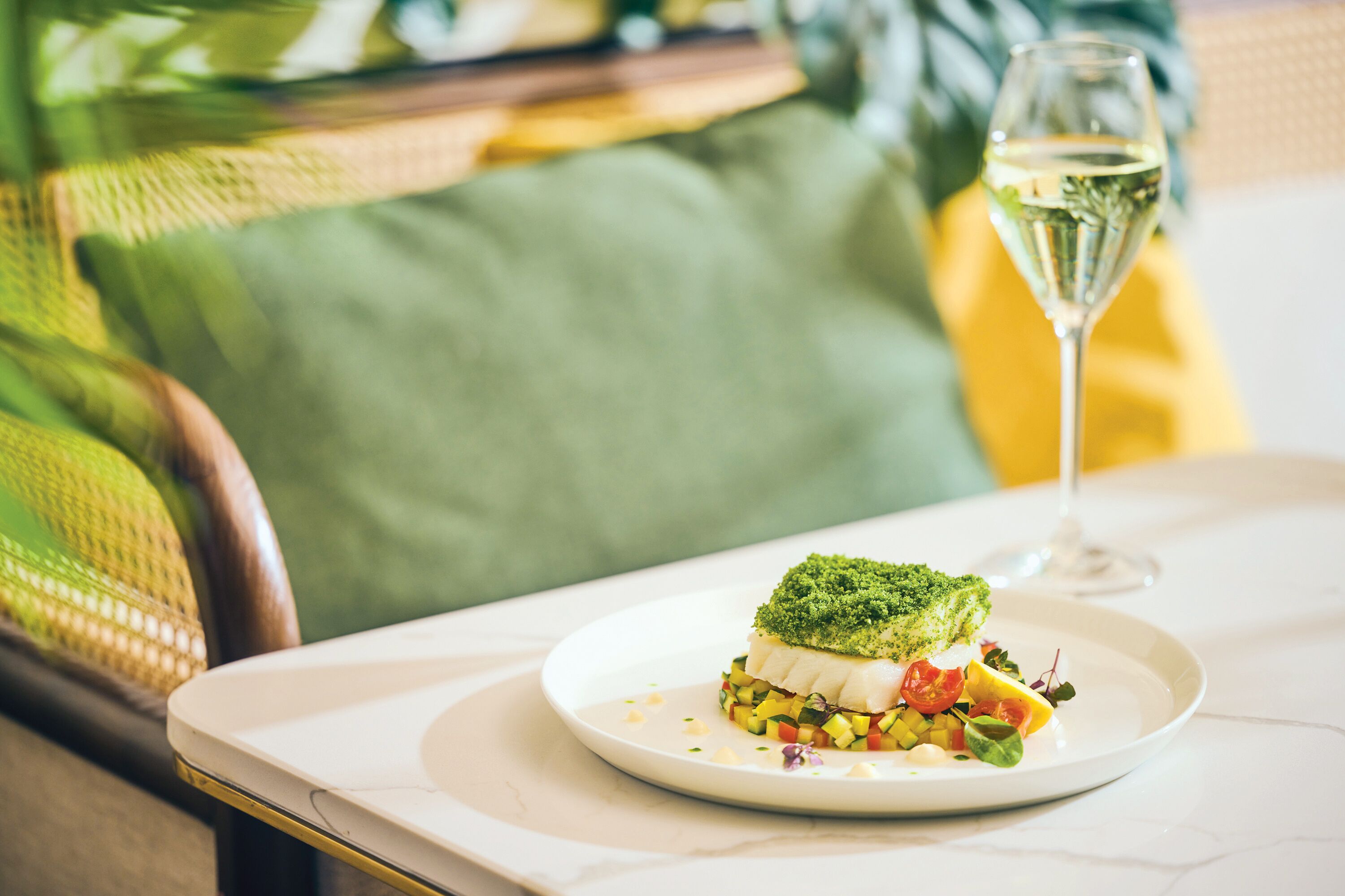 A plate with a dish on a table, a glass of white wine, and a green cushion in the background.