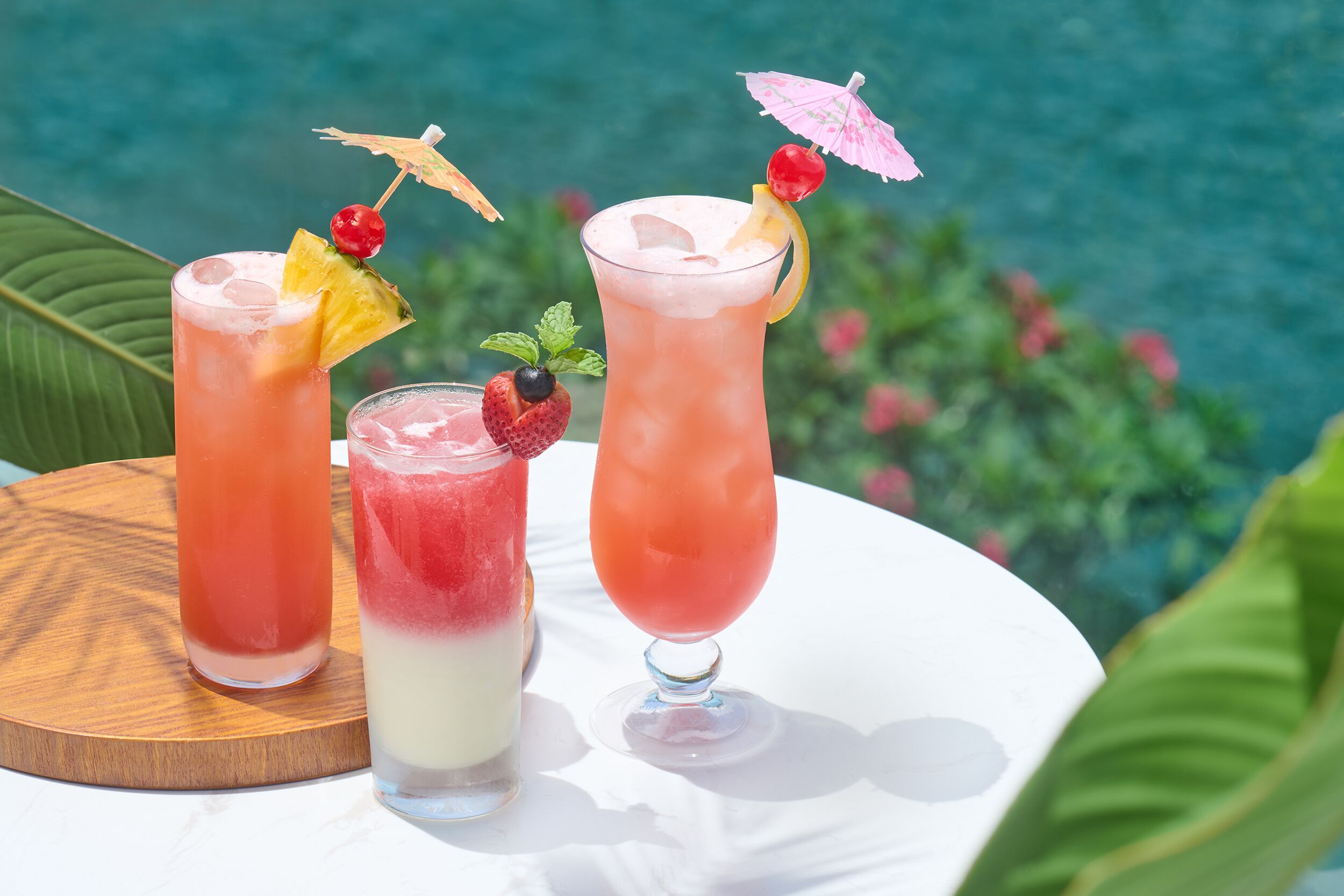 Three tropical drinks on a white table with a wooden coaster, set against a backdrop of a pool and greenery.