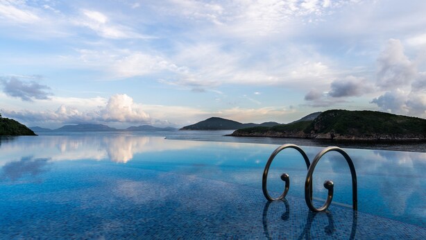 A clear blue infinity pool with metal railings, surrounded by calm water and distant mountains under a partly cloudy sky.