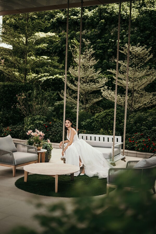 Une femme vêtue d’une robe blanche est assise sur une chaise suspendue dans un jardin. On y voit également un banc blanc et une table ronde ornée de fleurs.