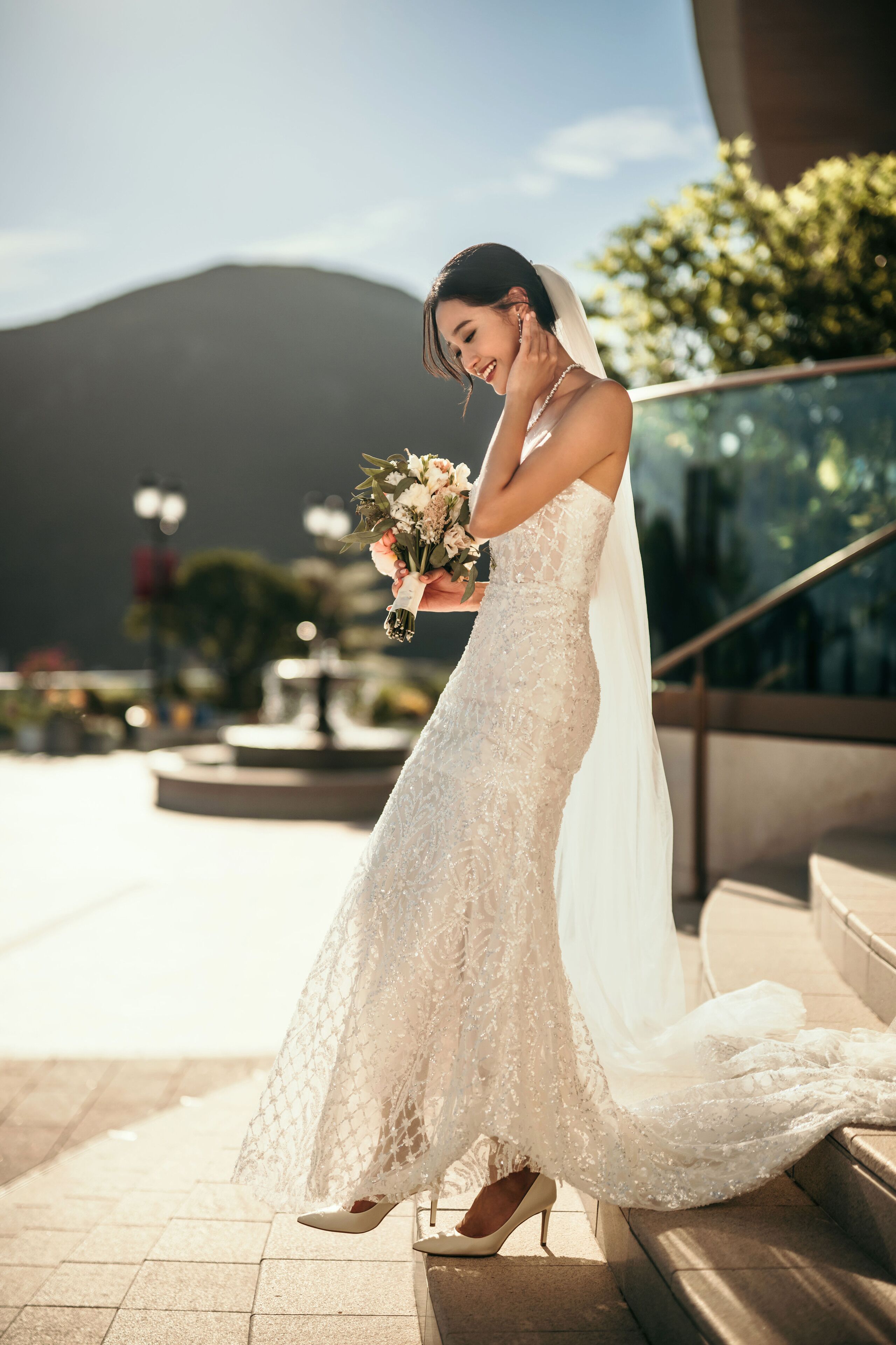 A bride in a white lace wedding dress holding a bouquet, standing on steps with a mountainous background.
