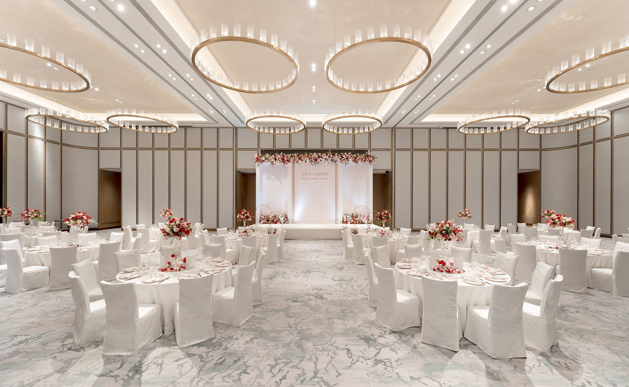 Um amplo salão de banquetes ricamente decorado, com mesas redondas preparadas para um evento formal, apresentando um palco com arranjos florais e uma placa com a inscrição "The Fullerton Ocean Park Hotel".