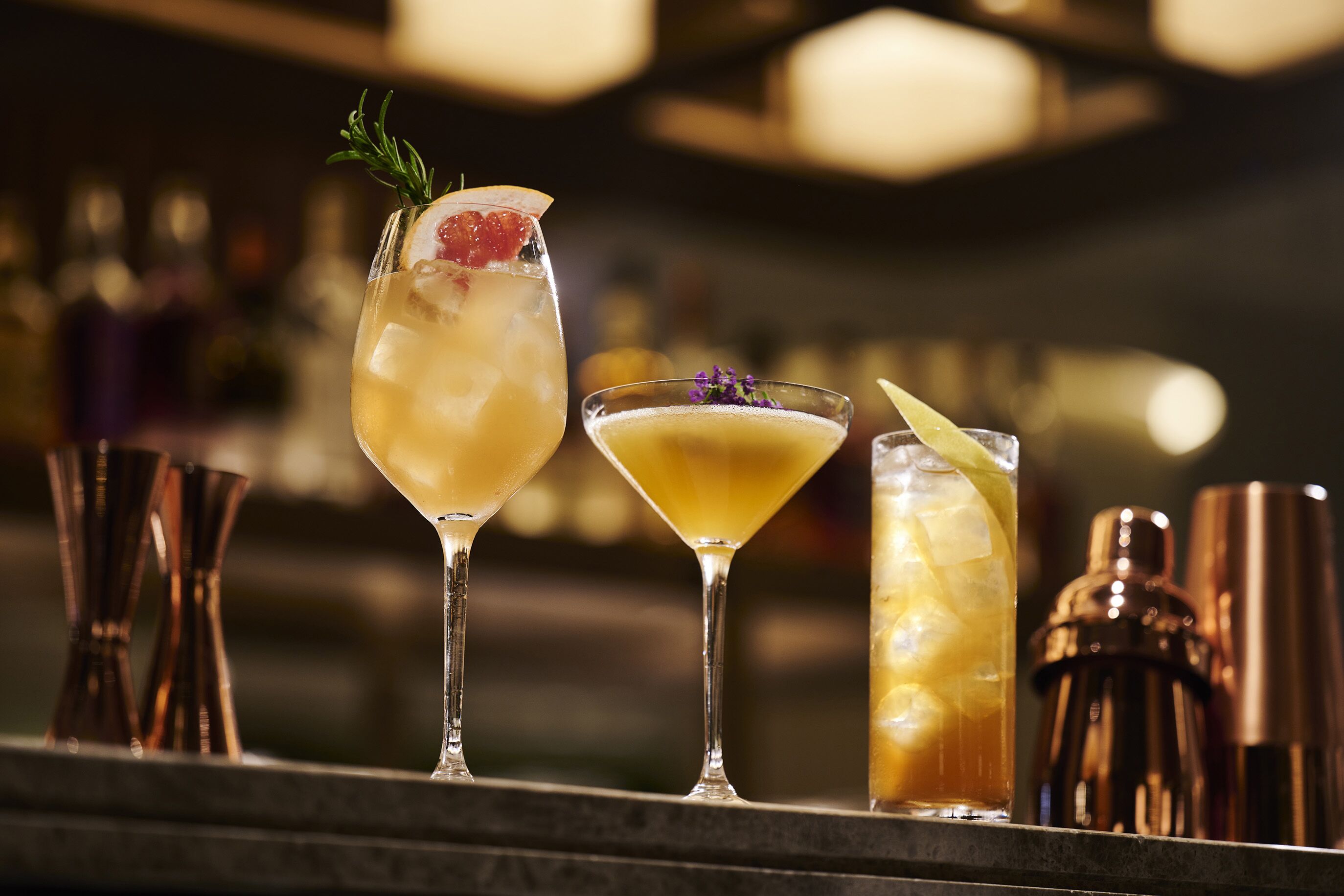 Three cocktails on a bar counter with copper shakers and bottles in the background.