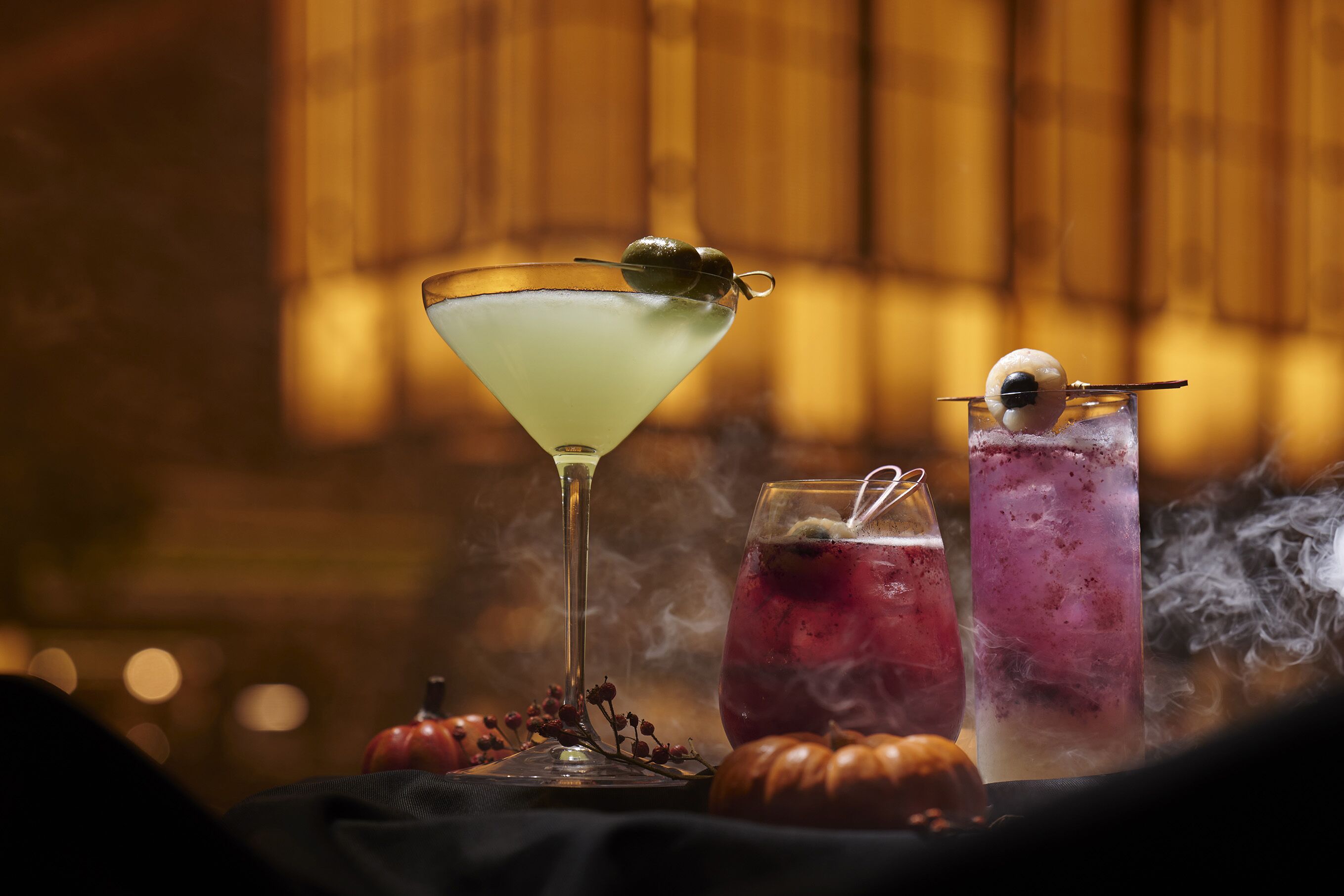 Un bicchiere Martini con guarnizione, un bicchiere alto con una cannuccia e un bicchiere basso con una cannuccia, il tutto su un tavolo con zucche e fumo.