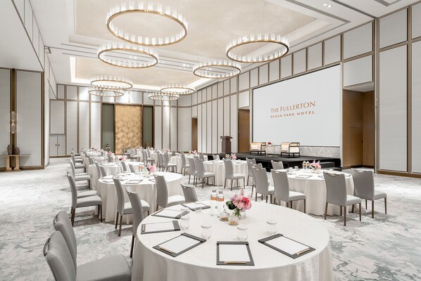 Ein Konferenzraum mit runden Tischen, Stühlen und einem großen Bildschirm, der "The Fullerton Ocean Park Hotel" zeigt.