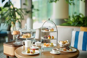 A three-tiered dessert stand with various pastries and desserts, a coffee pot, cups, and a teapot on a wooden table in a well-lit room with plants in the background. - The Fullerton Ocean Park Hotel Hong Kong (Hong Kong)