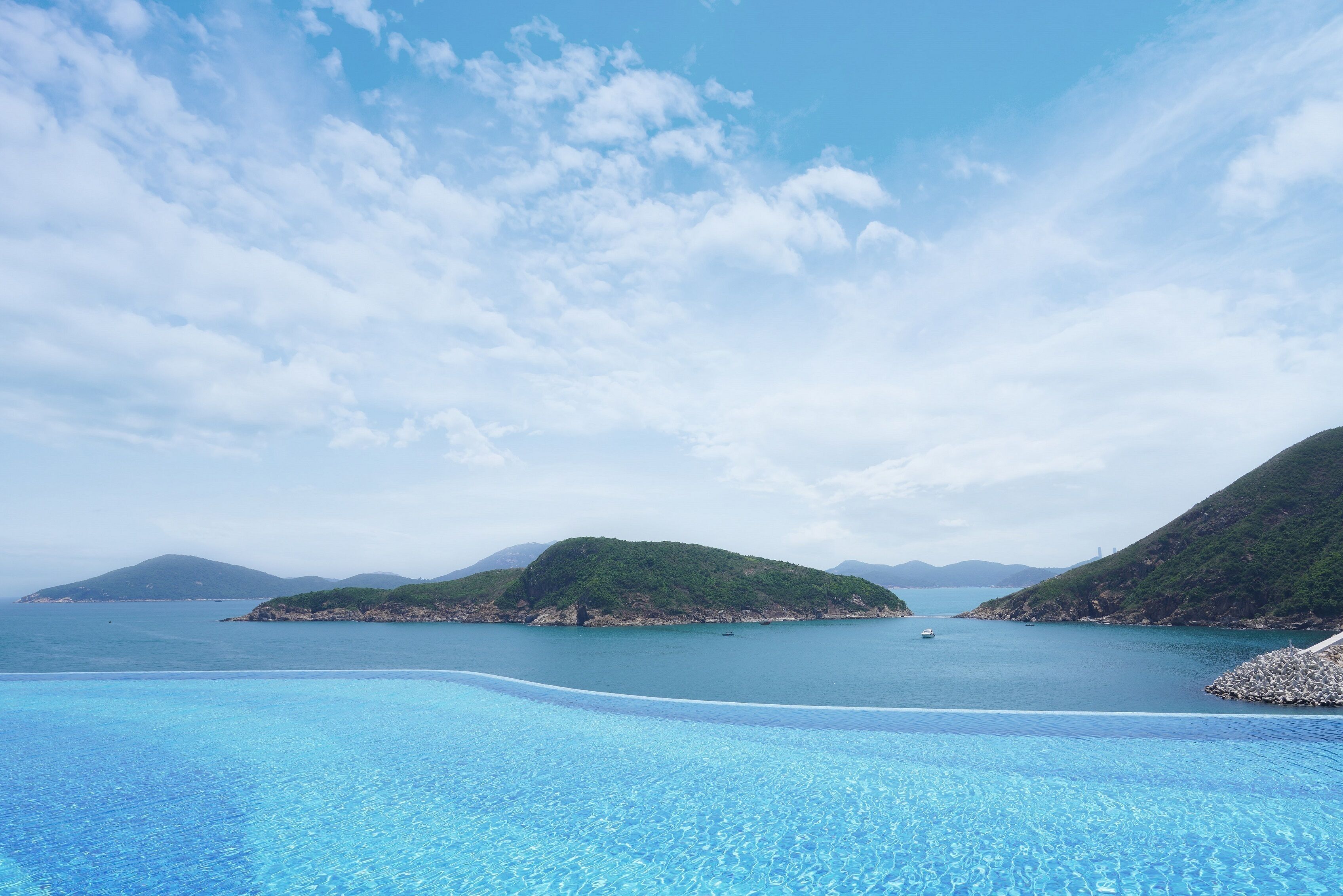 Une piscine d’un bleu éclatant, avec vue sur une mer calme et des collines verdoyantes en arrière-plan.