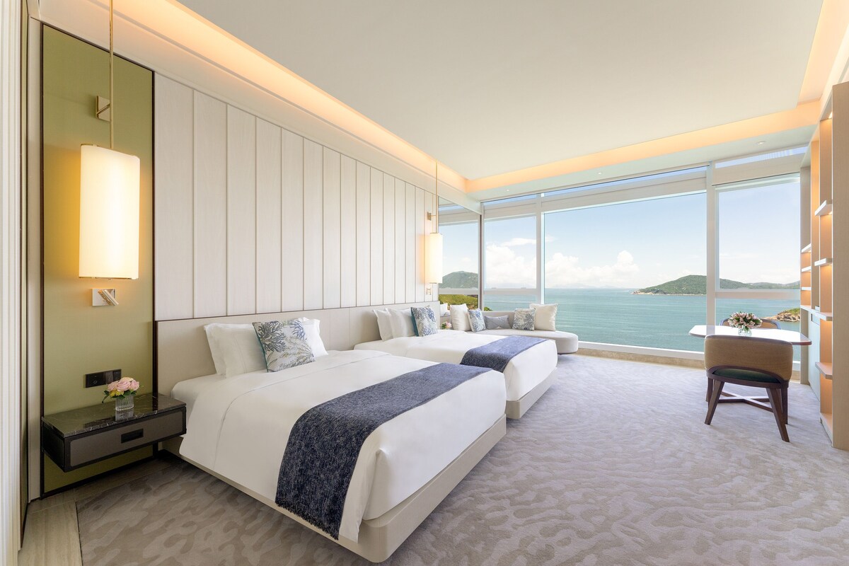 Une chambre d’hôtel moderne avec deux lits, une grande fenêtre donnant sur la mer et une petite table avec une chaise.
