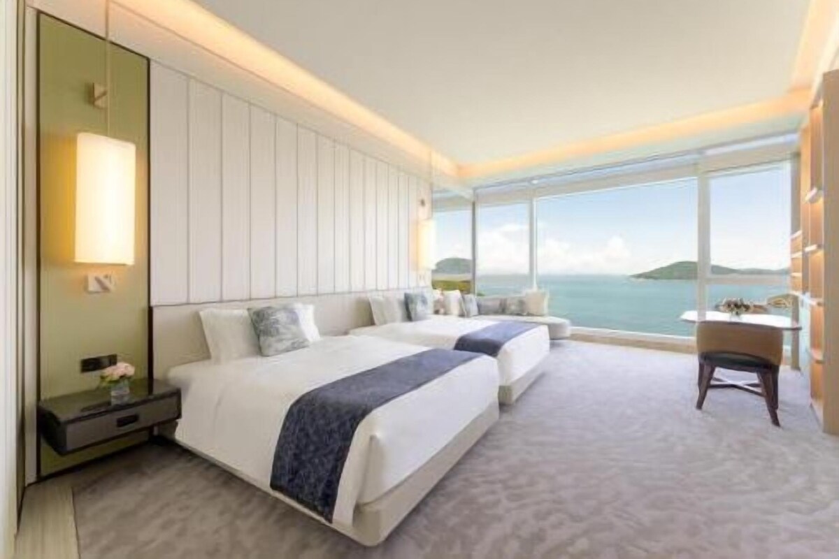 Une chambre d’hôtel avec deux lits, une grande fenêtre donnant sur la mer et une petite table avec une chaise.