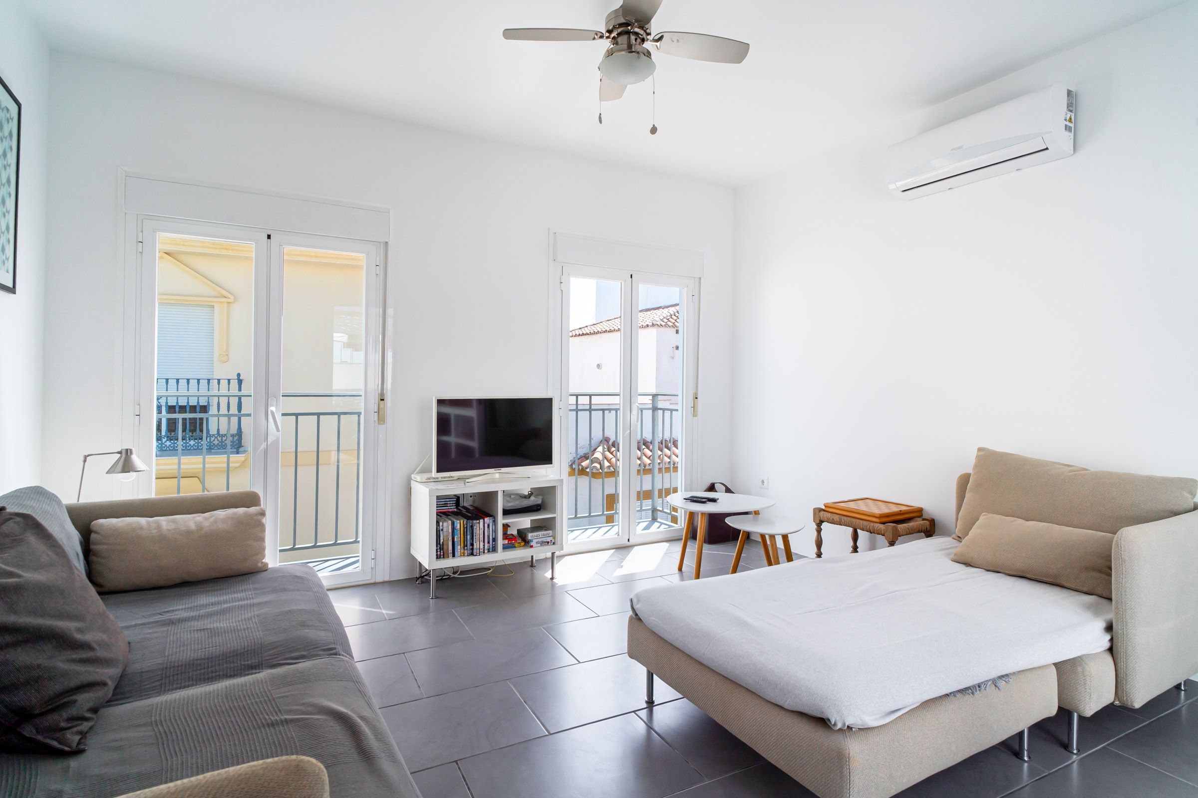 A modern living room with a sofa, a TV on a stand, a ceiling fan, and a balcony with a railing.