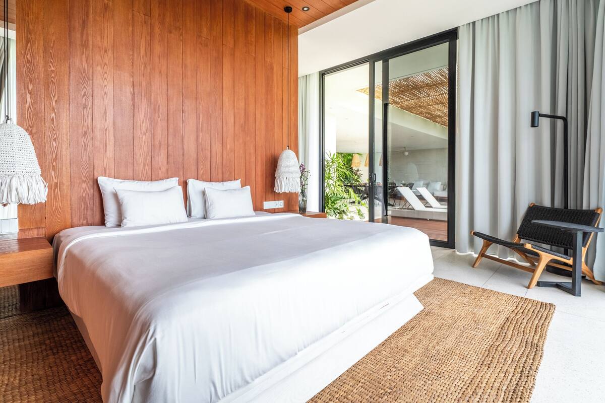 A modern bedroom with a large bed, wooden panel walls, a woven rug, and a view of a balcony with plants.