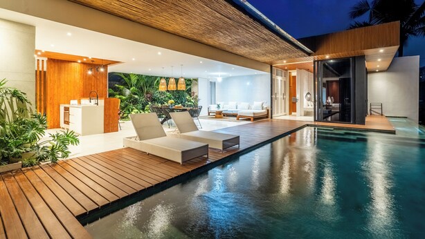 A modern outdoor pool area with wooden decking, lounge chairs, and a view of the interior. - Green Flow Villa 21 (Ubud)